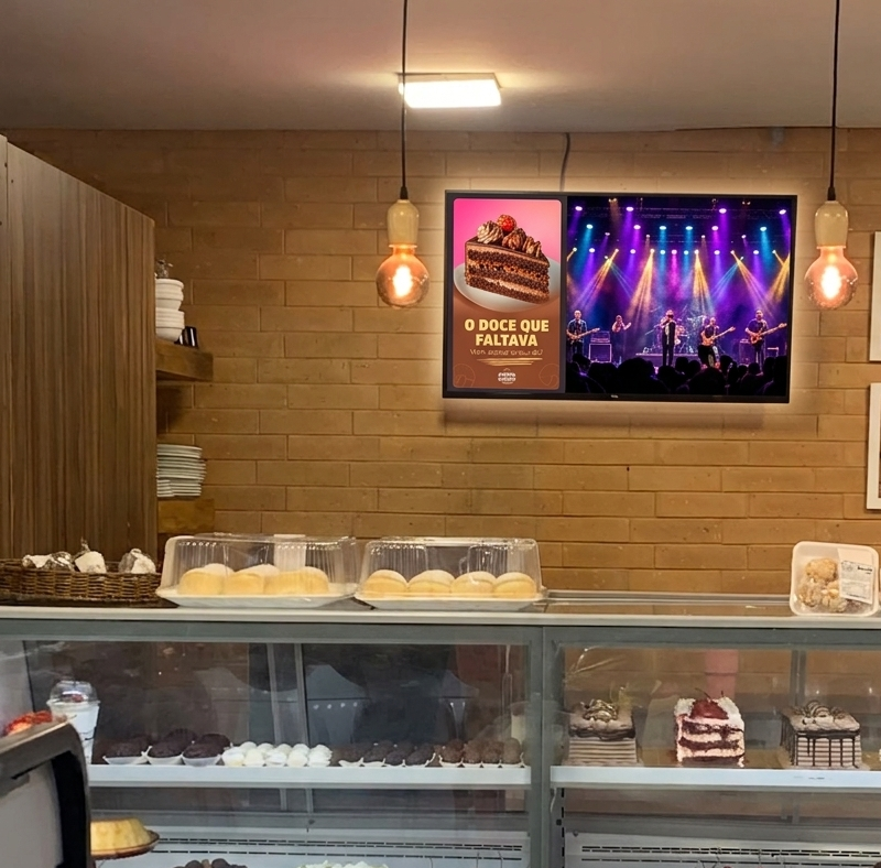 Balcão de padaria cheio de manhã, fila no caixa, e uma TV acima da vitrine mostrando um café e um pão de queijo quentinho.