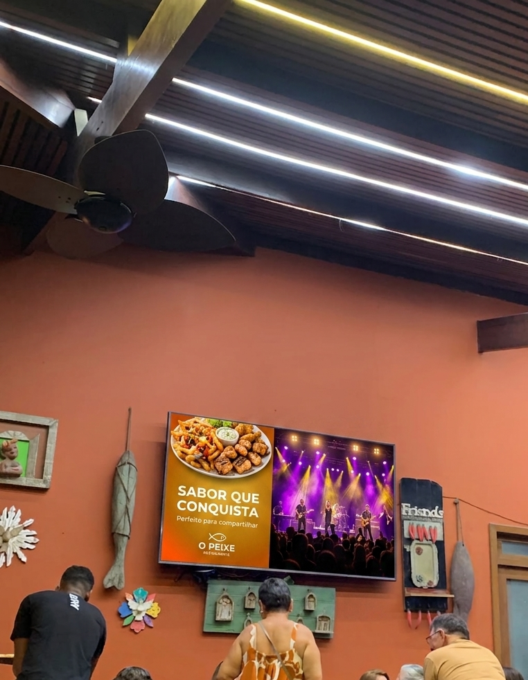 Entrada de bar movimentado à noite, pessoas esperando mesa em pé, uma telona mostrando o prato do dia e o aviso de um show no fim de semana.