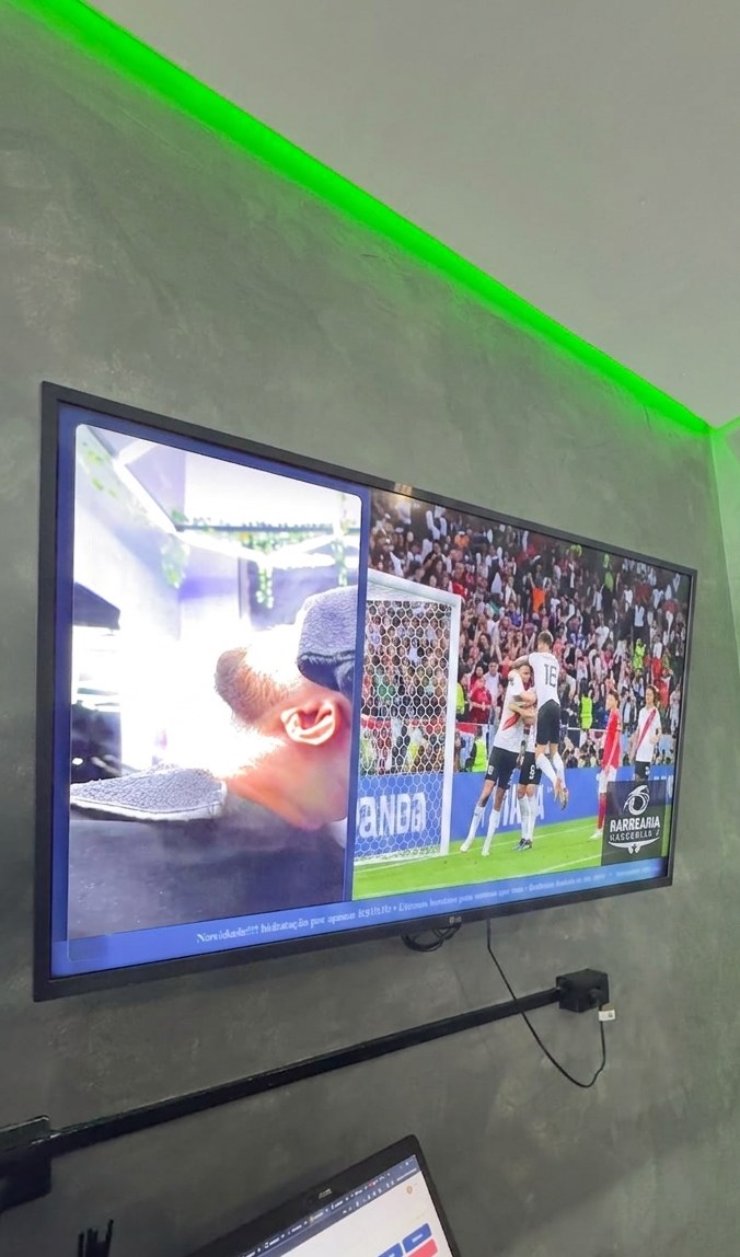 Cliente sentado na cadeira de barbeiro, de capa, olhando uma TV na parede que mostra a promoção de um combo barba + cabelo.